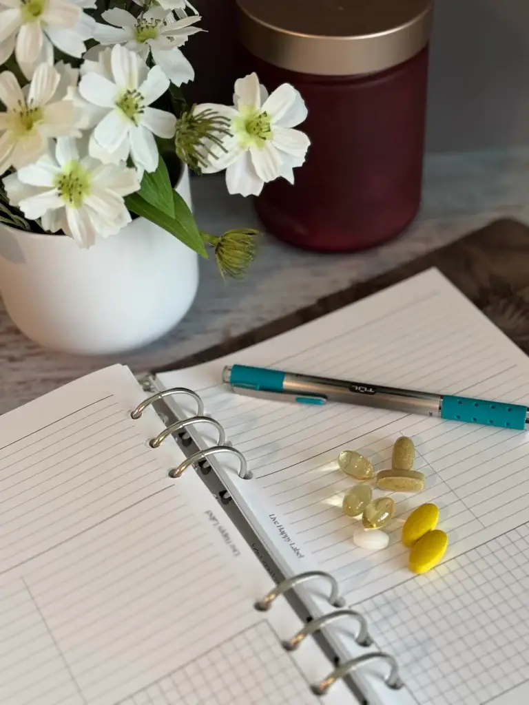 Open wellness planner with various daily supplements, a pen, and a small vase of white flowers on a desk.